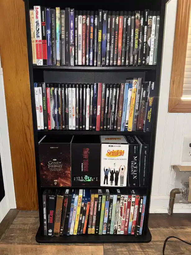 A black media shelf filled with numerous DVD and Blu-ray cases, including titles like 'Game of Thrones', 'Seinfeld', 'The…