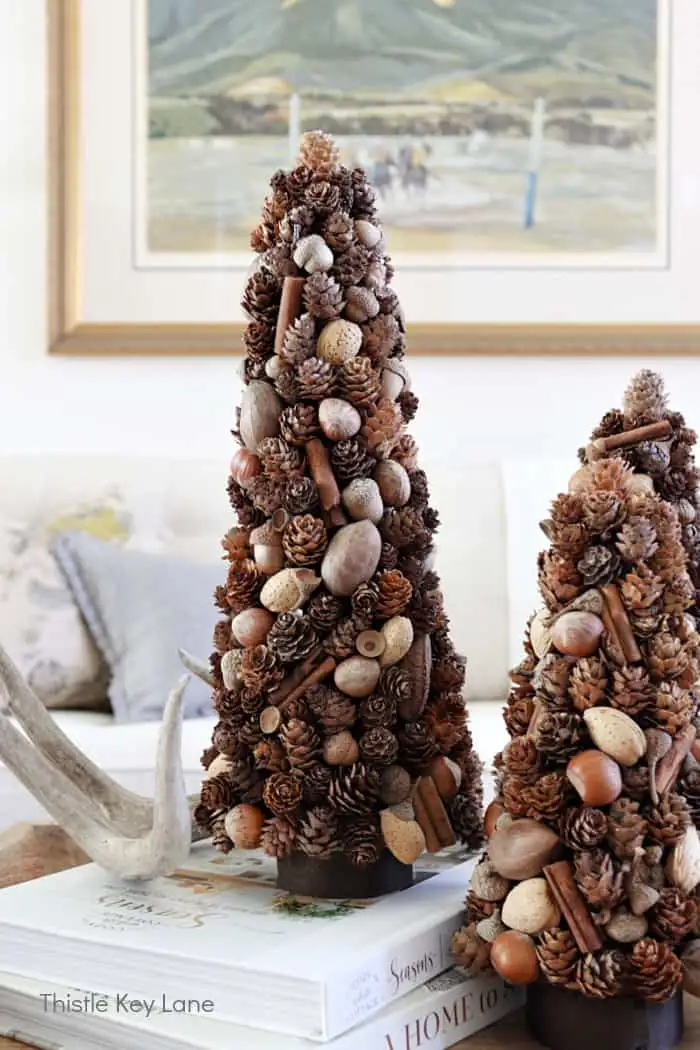 Two decorative, cone-shaped trees made from pinecones, acorns, and cinnamon sticks sit atop stacked books in a cozy living…