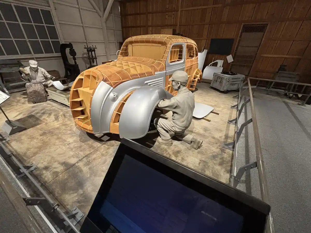 A museum diorama shows two life-sized statues of men working on a vintage car with a wooden frame and silver fenders. One…