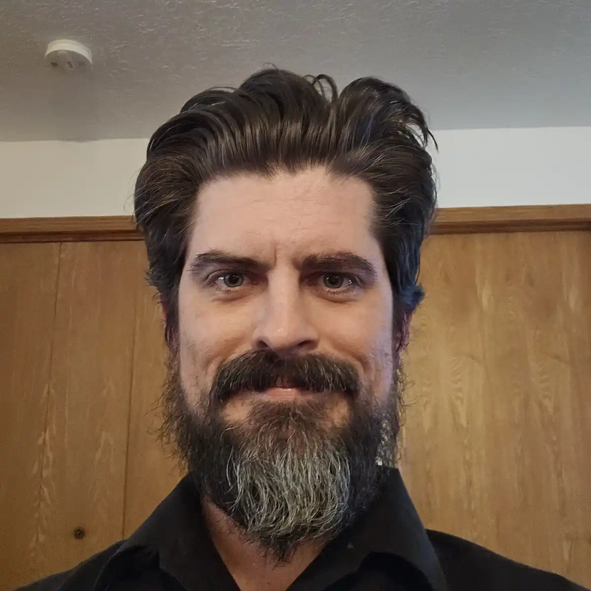 A close-up photo of a man with a full beard and mustache, showing gray hairs at the chin, wearing a black shirt against a…
