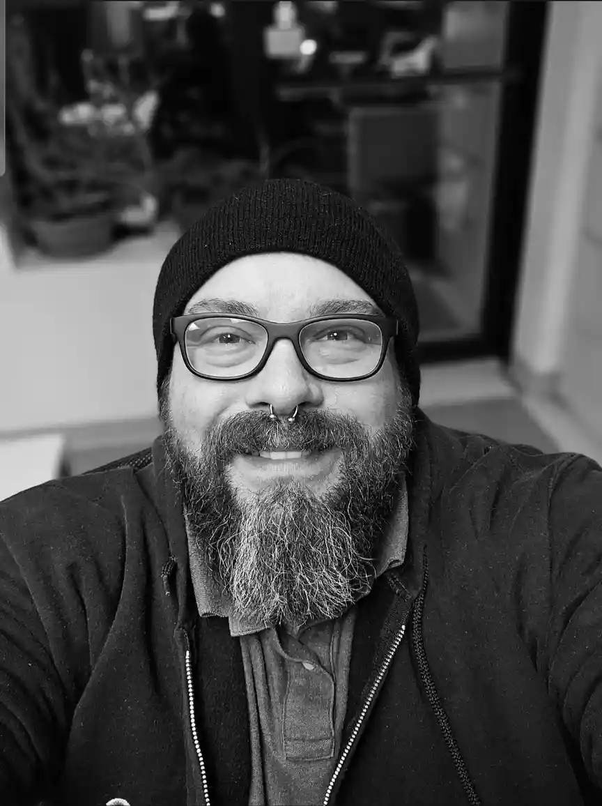 A black and white selfie of a smiling man with a full beard, glasses, and a beanie, looking directly at the camera. He has a…