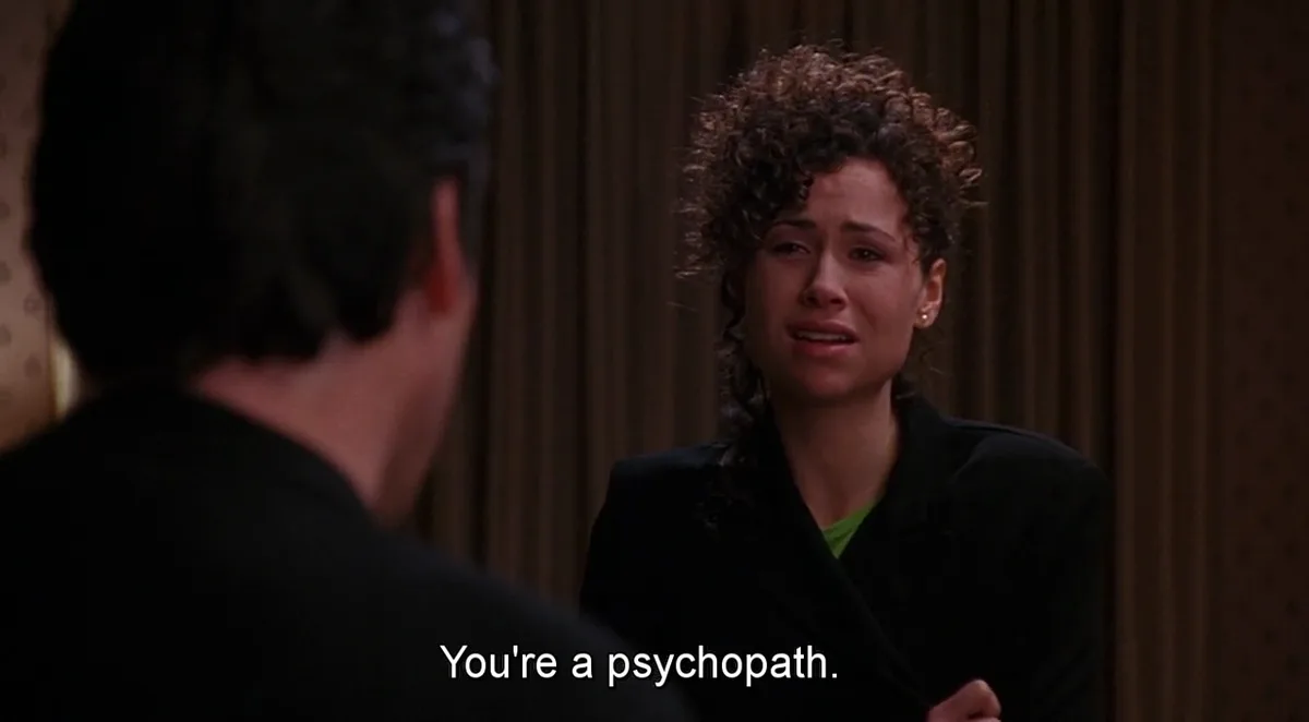 A woman with curly hair, wearing a black blazer over a green shirt, is visibly crying and looking distressed while facing a…