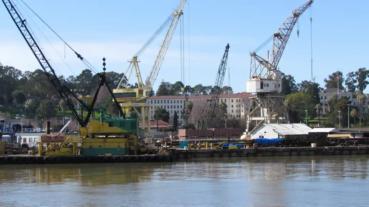 A wide shot of a harbor with multiple large industrial cranes, some yellow and some black, docked along a pier with water in…