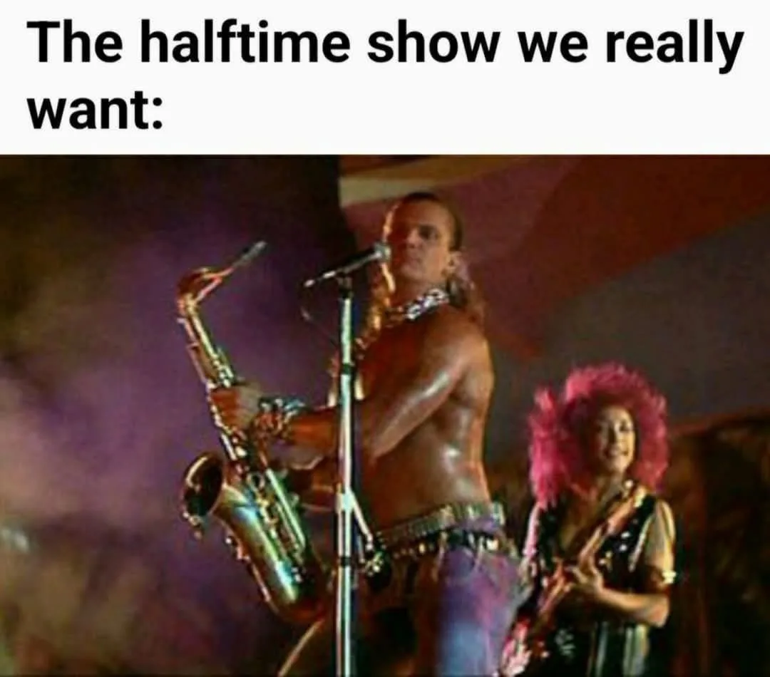 A shirtless man with long hair plays a saxophone on stage while a woman with a bright pink wig stands beside him holding a…