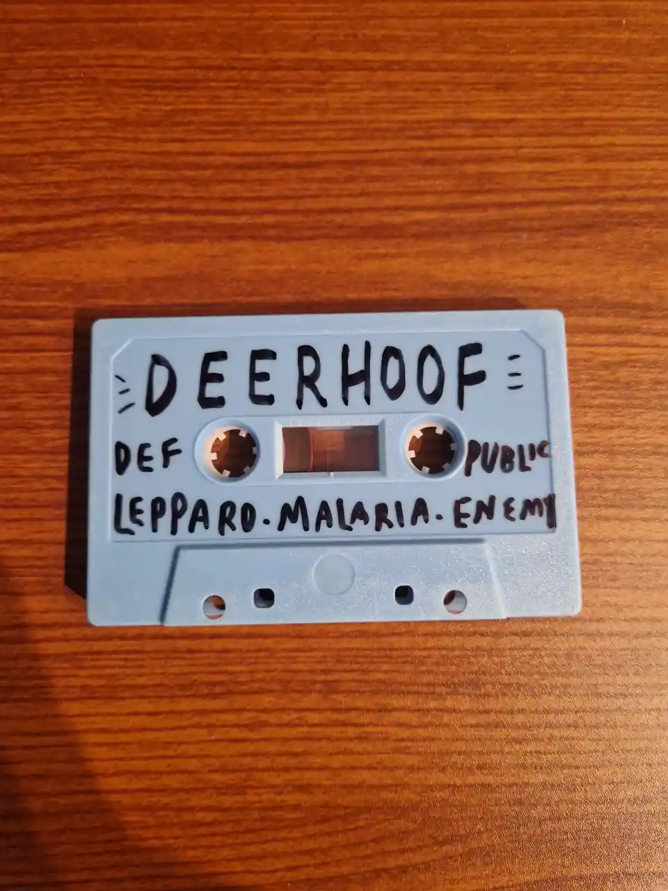 A light blue cassette tape sits on a wooden surface with handwritten text. The top line reads 'DEERHOOF' flanked by three…