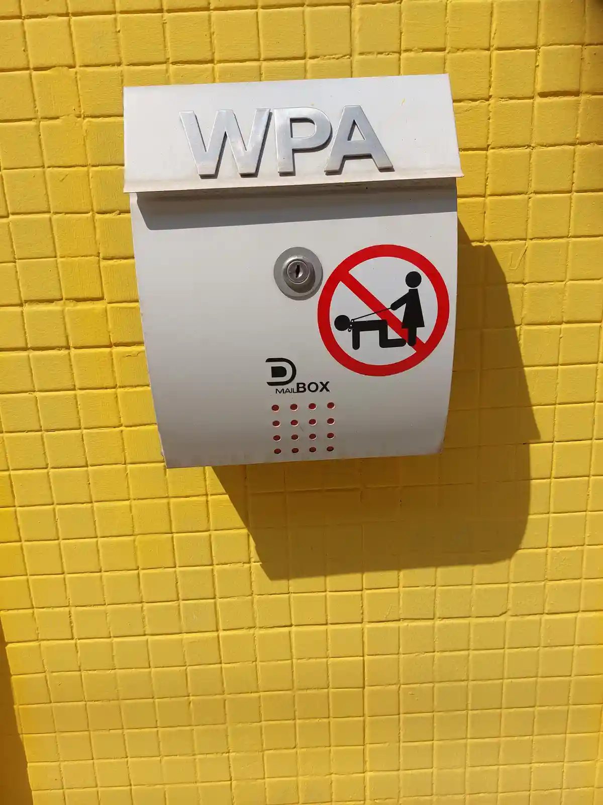 A white mailbox mounted on a yellow tiled wall has the letters 'WPA' on its top. It features a 'no' symbol over a pictogram…