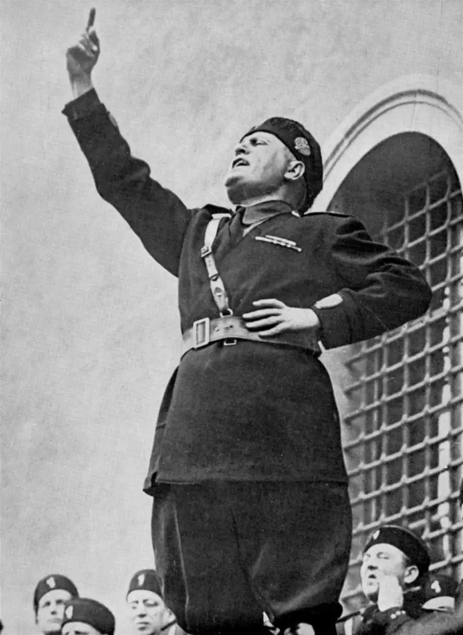 A black-and-white historical photo shows a man in a military-style uniform, raising his right arm with index finger pointing…