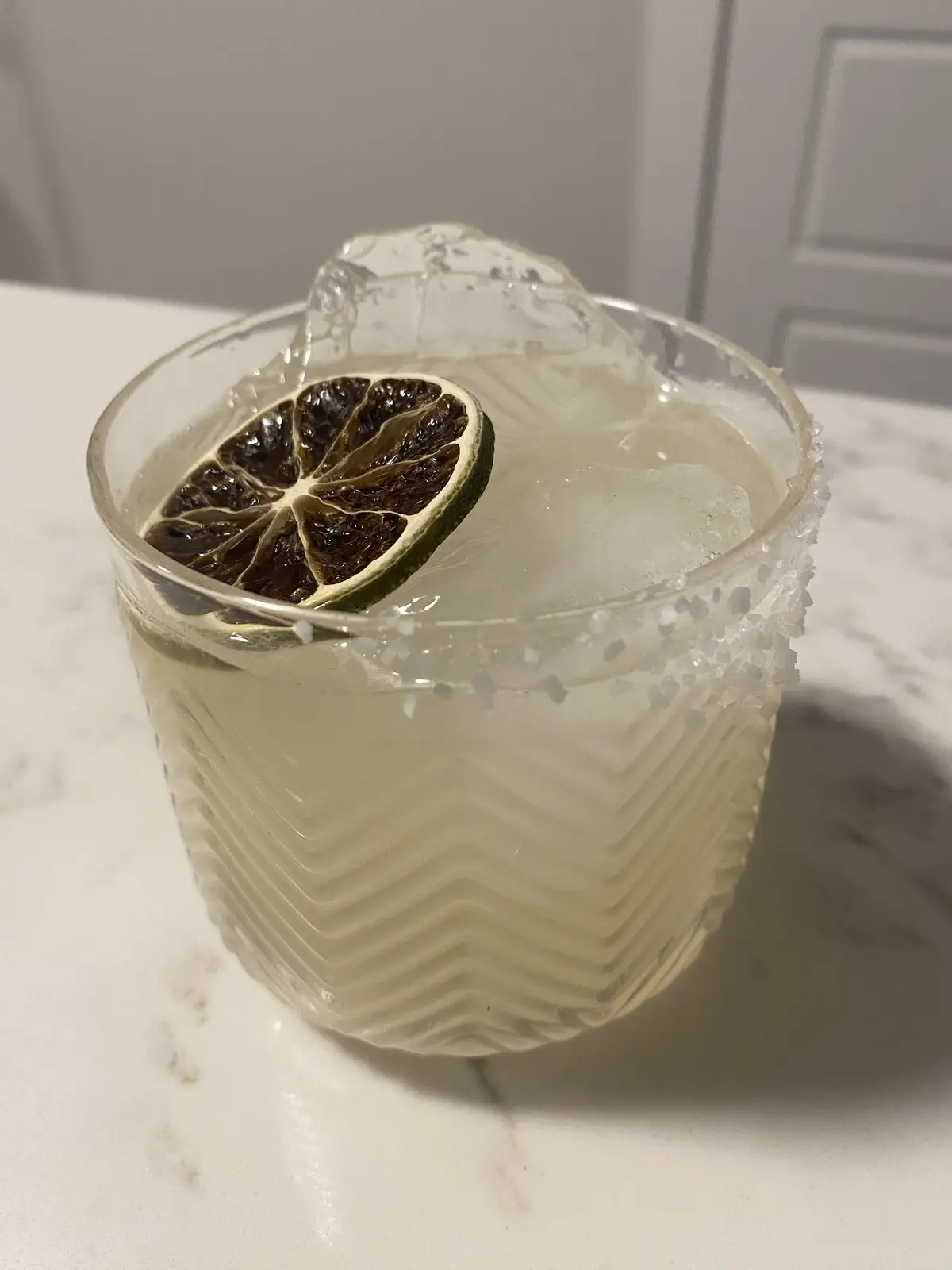 A close-up photo shows a cocktail in a textured glass with a salted rim and a charred lime wheel garnish, sitting on a white…