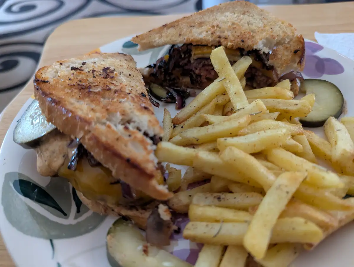 A close-up photo shows a toasted sandwich cut in half, served with golden fries and pickles on a patterned plate. The…