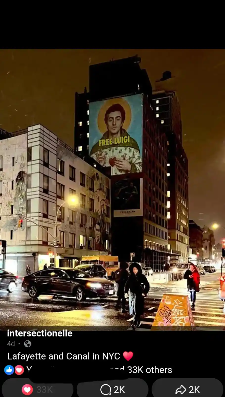 A nighttime street scene in New York City shows pedestrians crossing a wet street with cars passing by. A large billboard on…