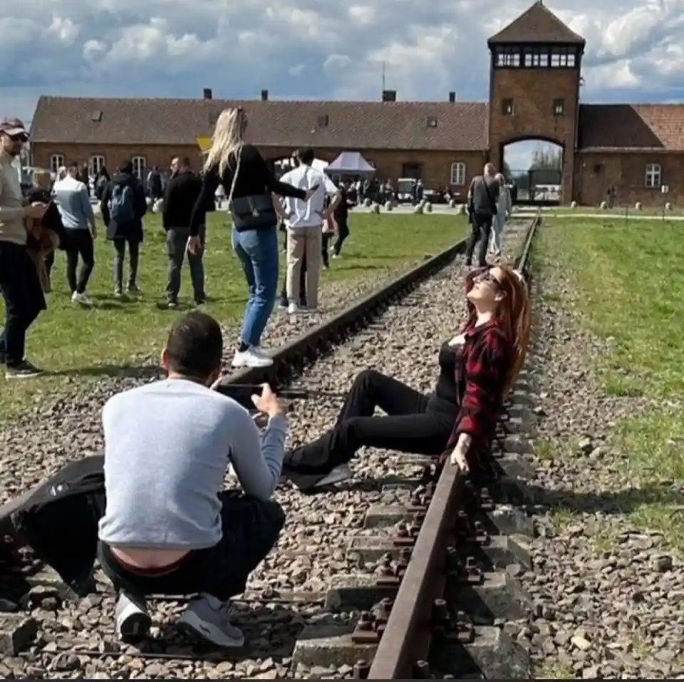 A woman with red hair and sunglasses sits casually on train tracks at what appears to be Auschwitz-Birkenau, with a man…