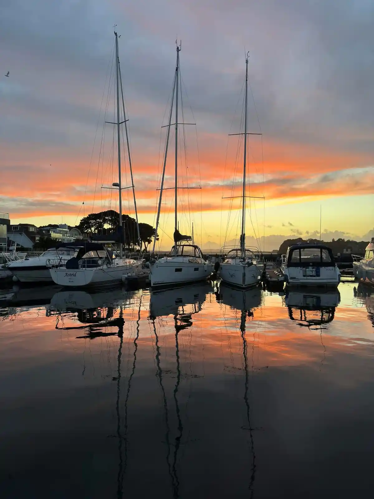 A serene marina at sunset, featuring several sailboats docked in calm water that perfectly mirrors the vibrant orange and…