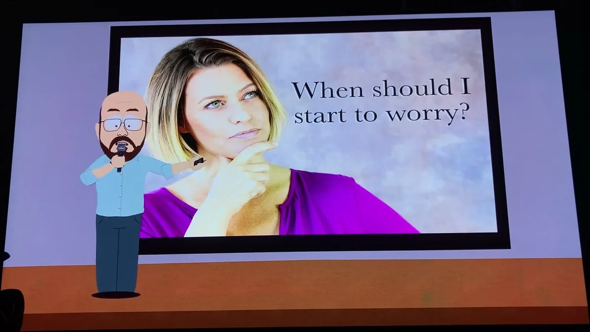 A cartoon man with glasses and a beard stands on a stage holding a microphone, positioned in front of a large screen. The…