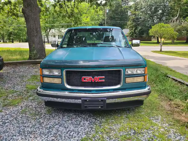 A teal GMC pickup truck is parked on a gravel driveway with grass growing through it, facing the camera. The vehicle has a…
