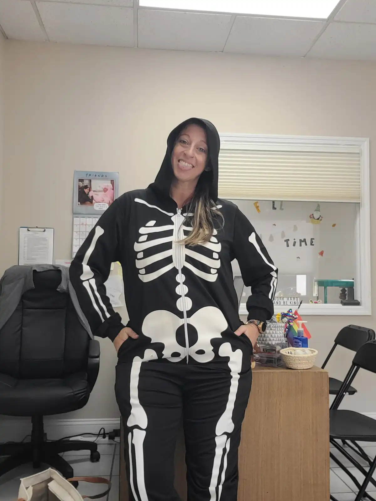 A woman in a black skeleton-themed hoodie and pants with white bone patterns stands smiling in an office, hands in pockets…