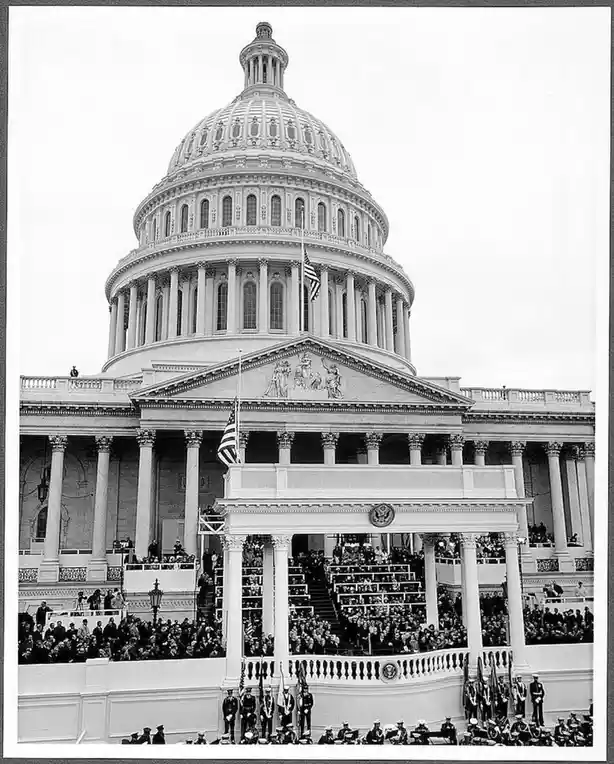 A black-and-white historical photograph shows the U.S. Capitol building with a large crowd gathered on its steps and in the…