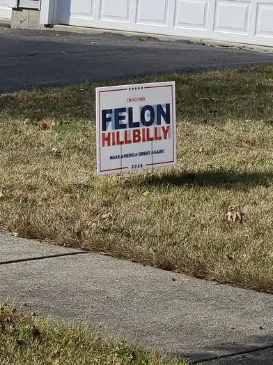 A political yard sign in a grassy lawn in front of a house with a white garage door. The sign, styled like a campaign poster…