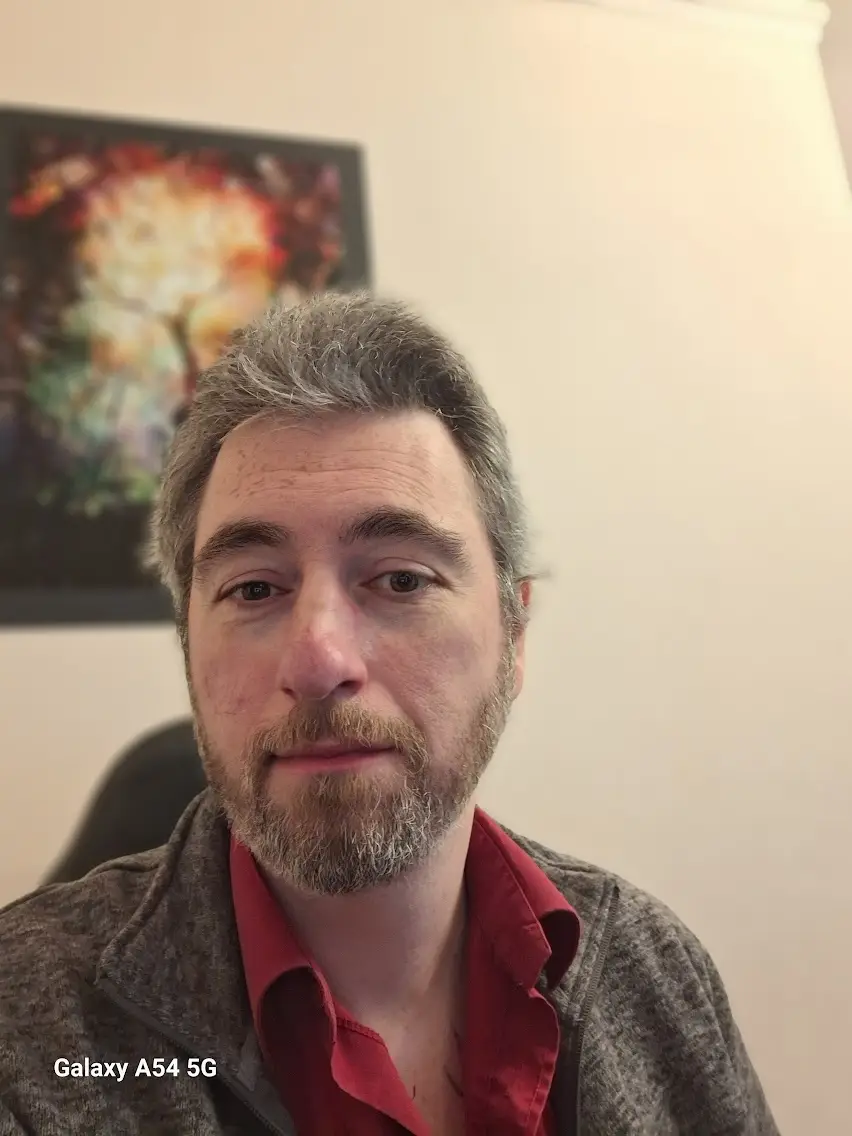 A close-up selfie of a middle-aged man with graying hair and a beard, wearing a red shirt under a gray jacket, looking…