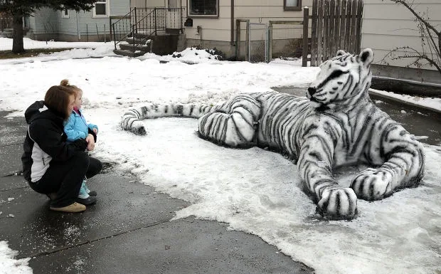 A woman holding a small child crouches in a snowy driveway, looking at a large, intricate snow sculpture shaped like a white…
