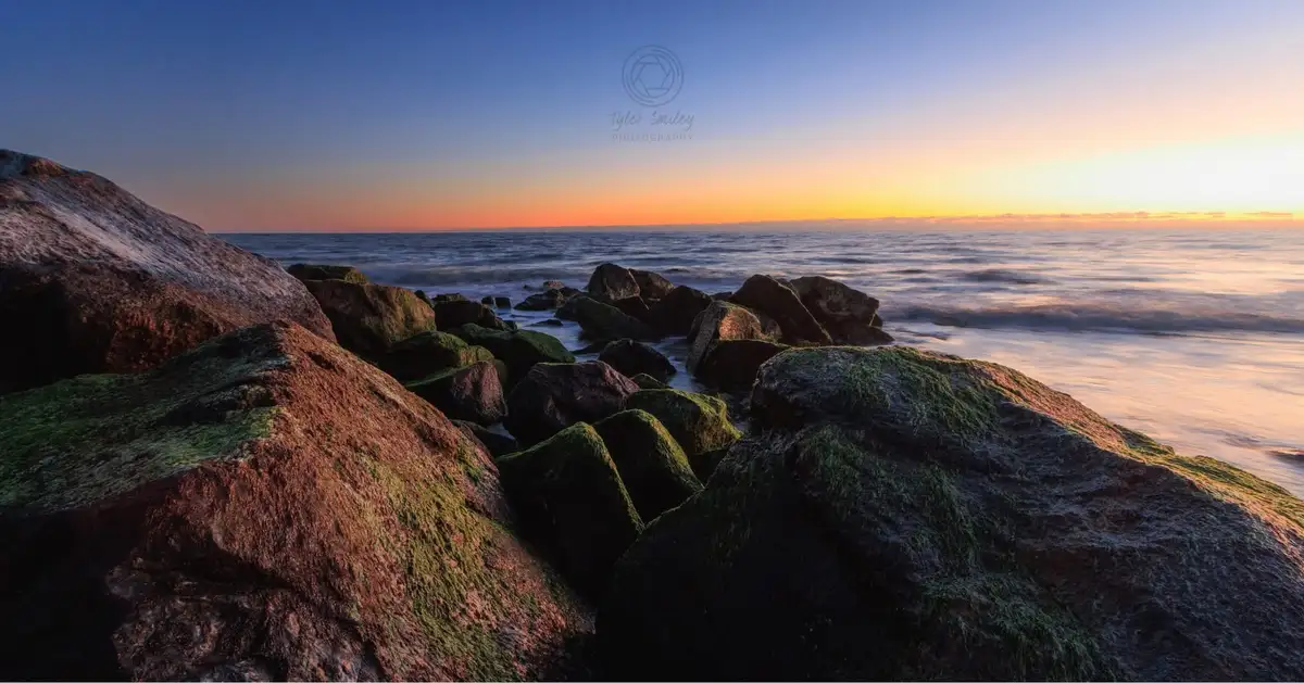 A serene landscape photo of moss-covered rocks at sunset, with calm water and a gradient sky from blue to orange. The image…