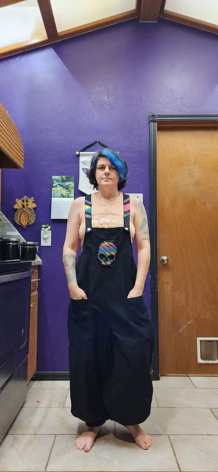 A person with blue-dyed hair and tattoos stands barefoot in a kitchen wearing black overalls over a tank top, hands in…