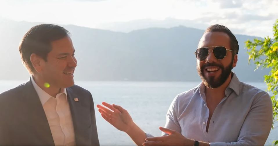 two men are smiling and talking outdoors near water with mountains in the background. The man on the right, wearing…