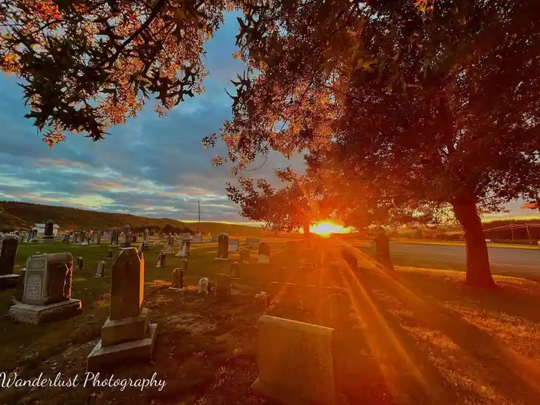 A serene cemetery at sunset, with numerous gravestones scattered across a grassy field under a dramatic sky. The sun is…