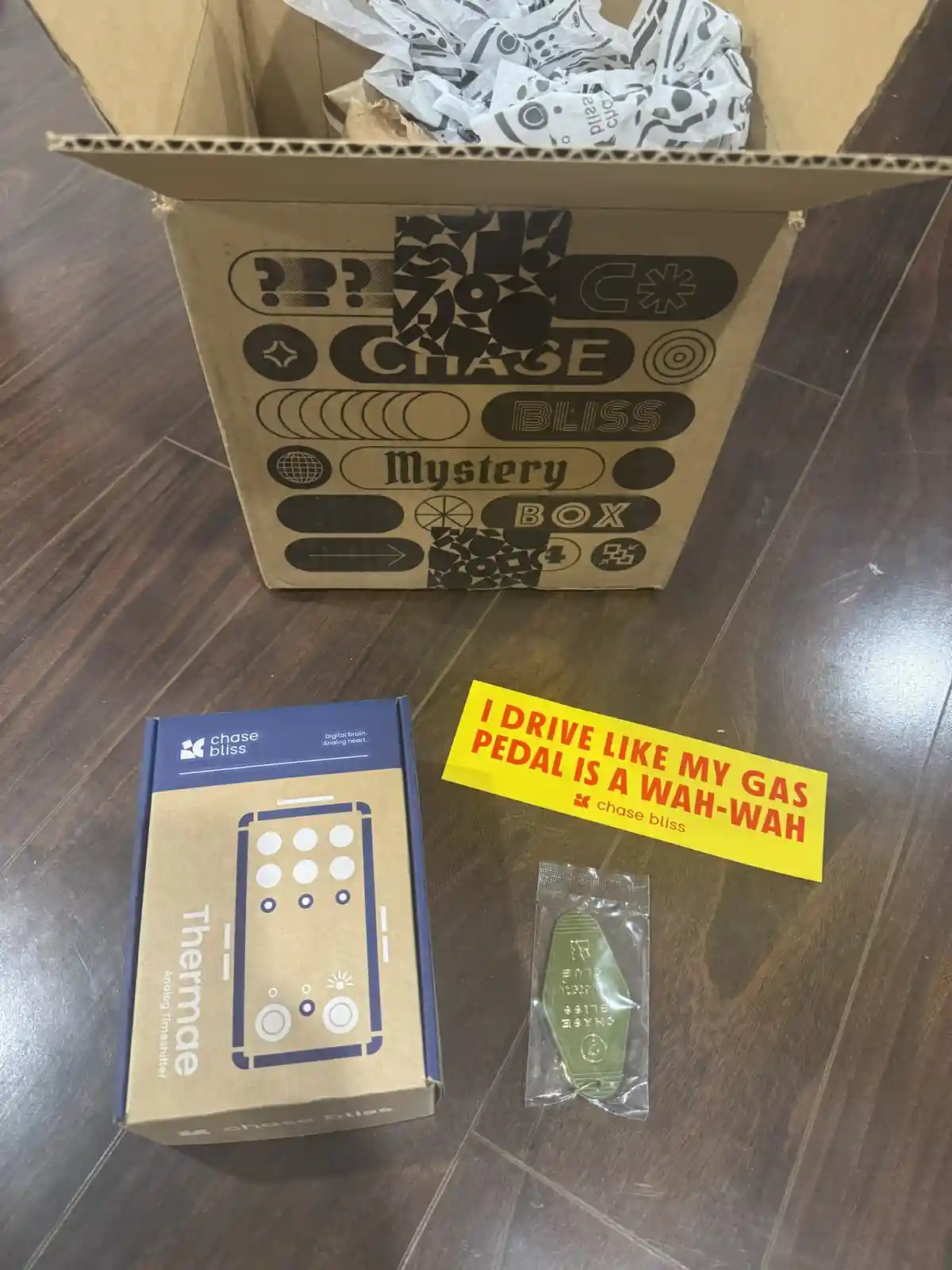 An open cardboard box labeled 'CHASE BLISS Mystery Box' sits on a wooden floor, filled with patterned packing paper. In…
