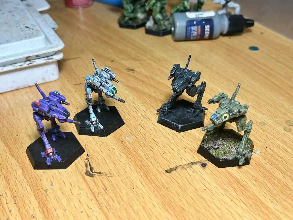 Four painted miniature mechs on hexagonal bases sit on a wooden table, each with distinct color schemes and paint jobs. In…