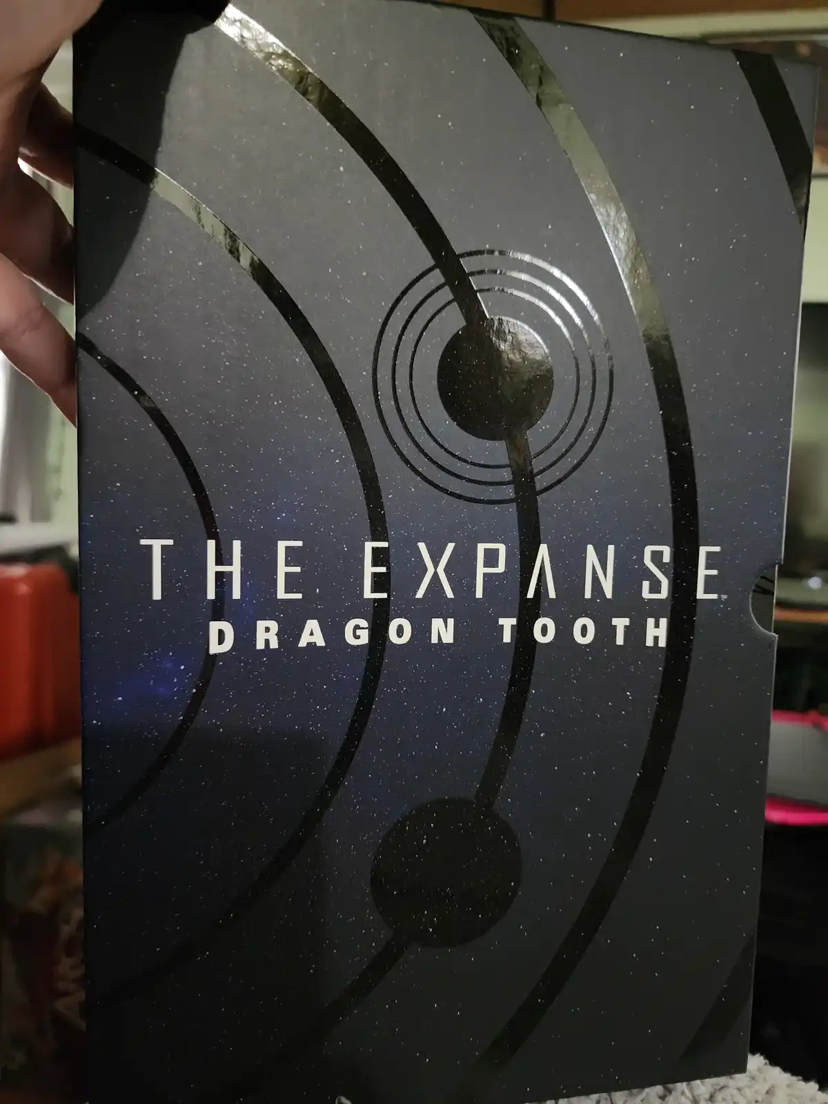 A hand holds a dark, space-themed box featuring a cosmic design with orbiting planets and stars. The text 'THE EXPANSE…