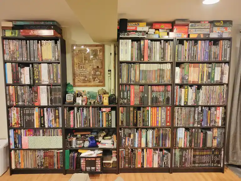 A room filled with tall, dark wooden bookshelves packed with board games and role-playing game books, with a framed map…