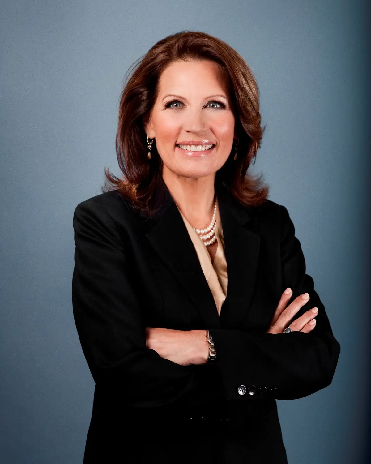 A professional headshot of a smiling woman with brown hair, wearing a black blazer over a beige blouse, a pearl necklace…