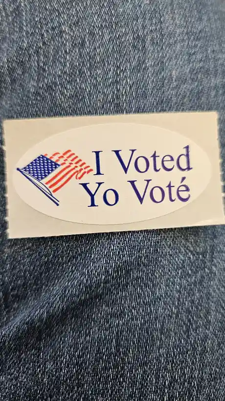 A close-up photo shows an oval 'I Voted' sticker with an American flag graphic, placed on blue denim fabric. The sticker's…