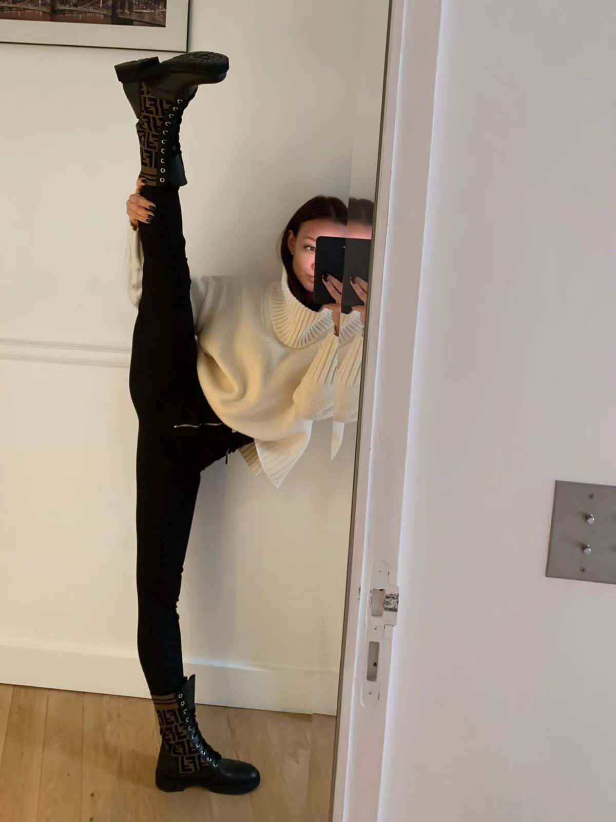 A person is taking a mirror selfie while holding one leg up high, wearing black Fendi boots and black leggings with a…