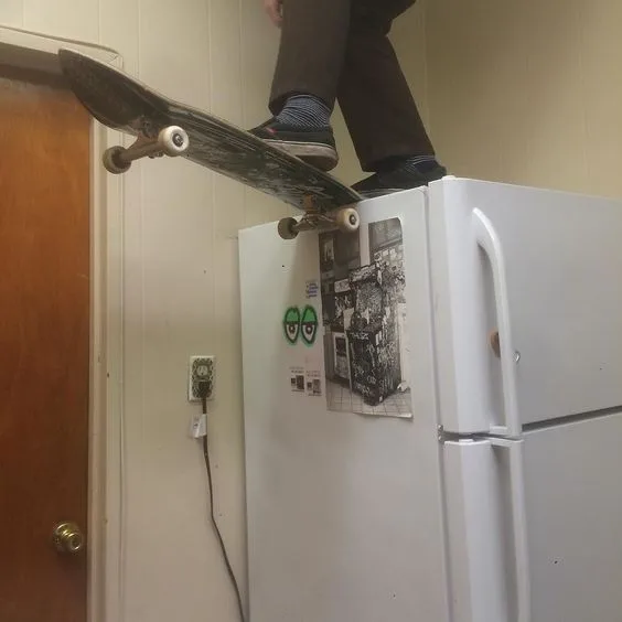 A person stands on top of a white refrigerator, balancing a skateboard on its edge with their feet. The fridge has magnets…