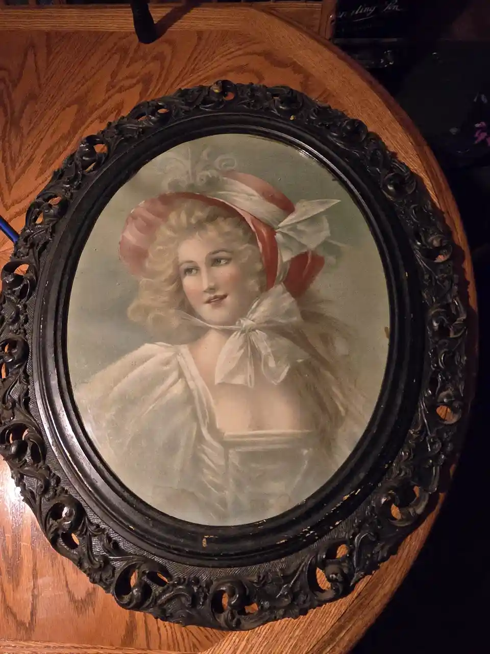 An oval portrait of a woman with curly blonde hair, wearing a red and white bonnet with a large bow, is framed in an ornate…