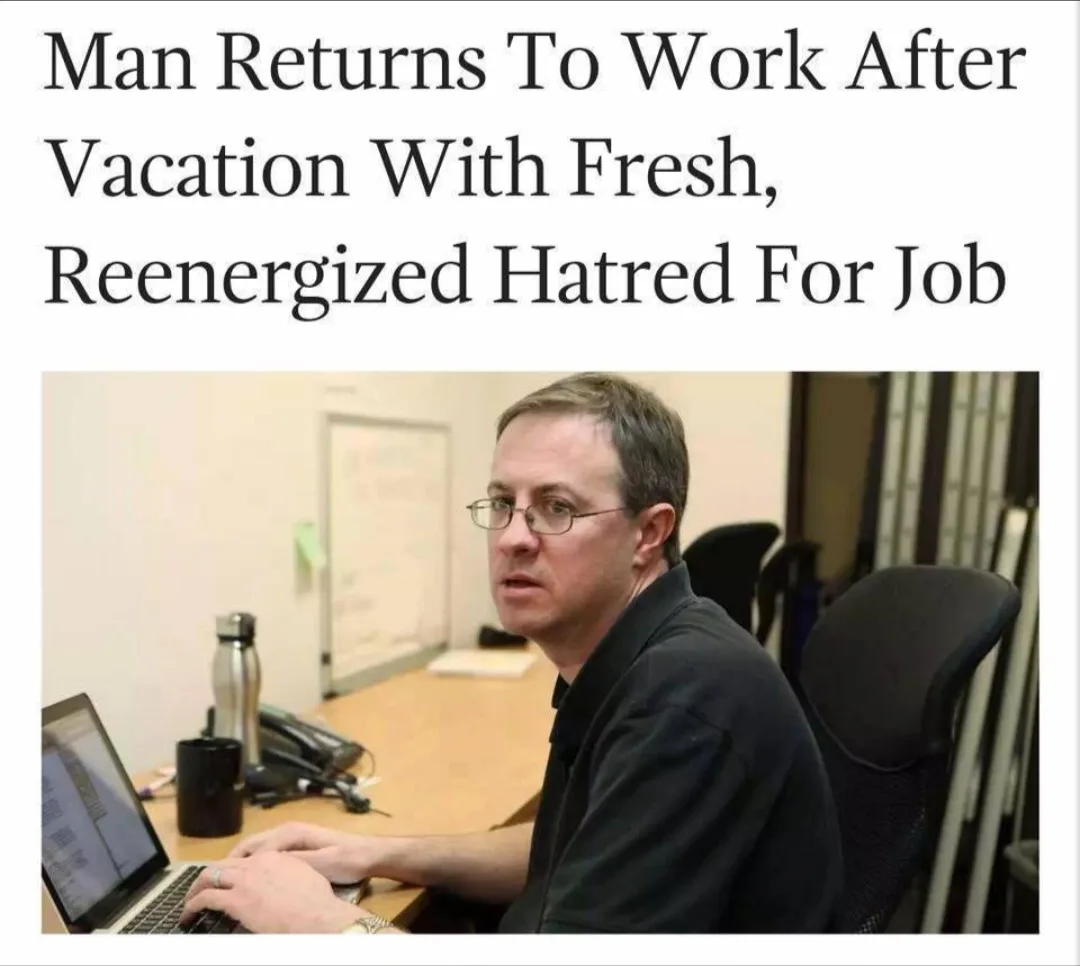 A man with glasses sits at a desk in an office, typing on a laptop while looking over his shoulder with a weary, annoyed…