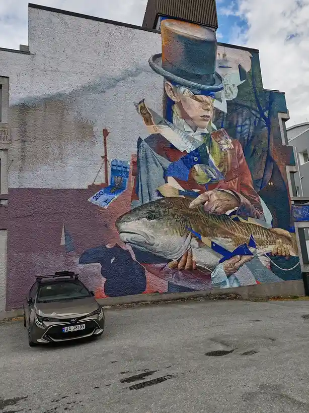 A silver Toyota Corolla with a roof rack is parked in front of a large mural on a building wall. The mural depicts a person…