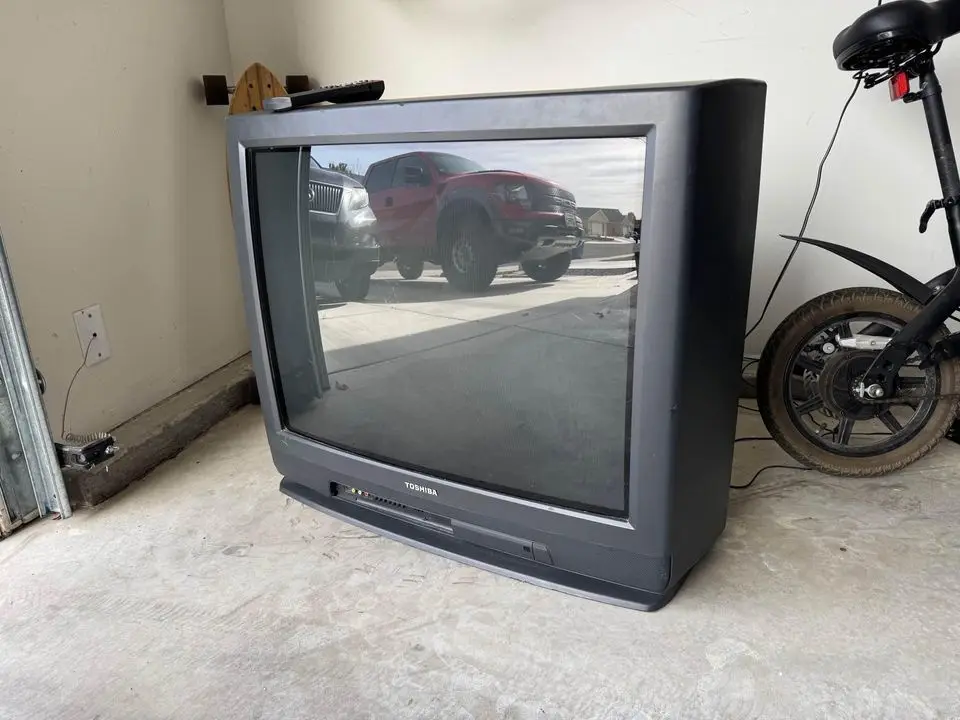 An old, large, black Toshiba CRT television sits unplugged on a concrete floor in a garage, its screen reflecting parked…
