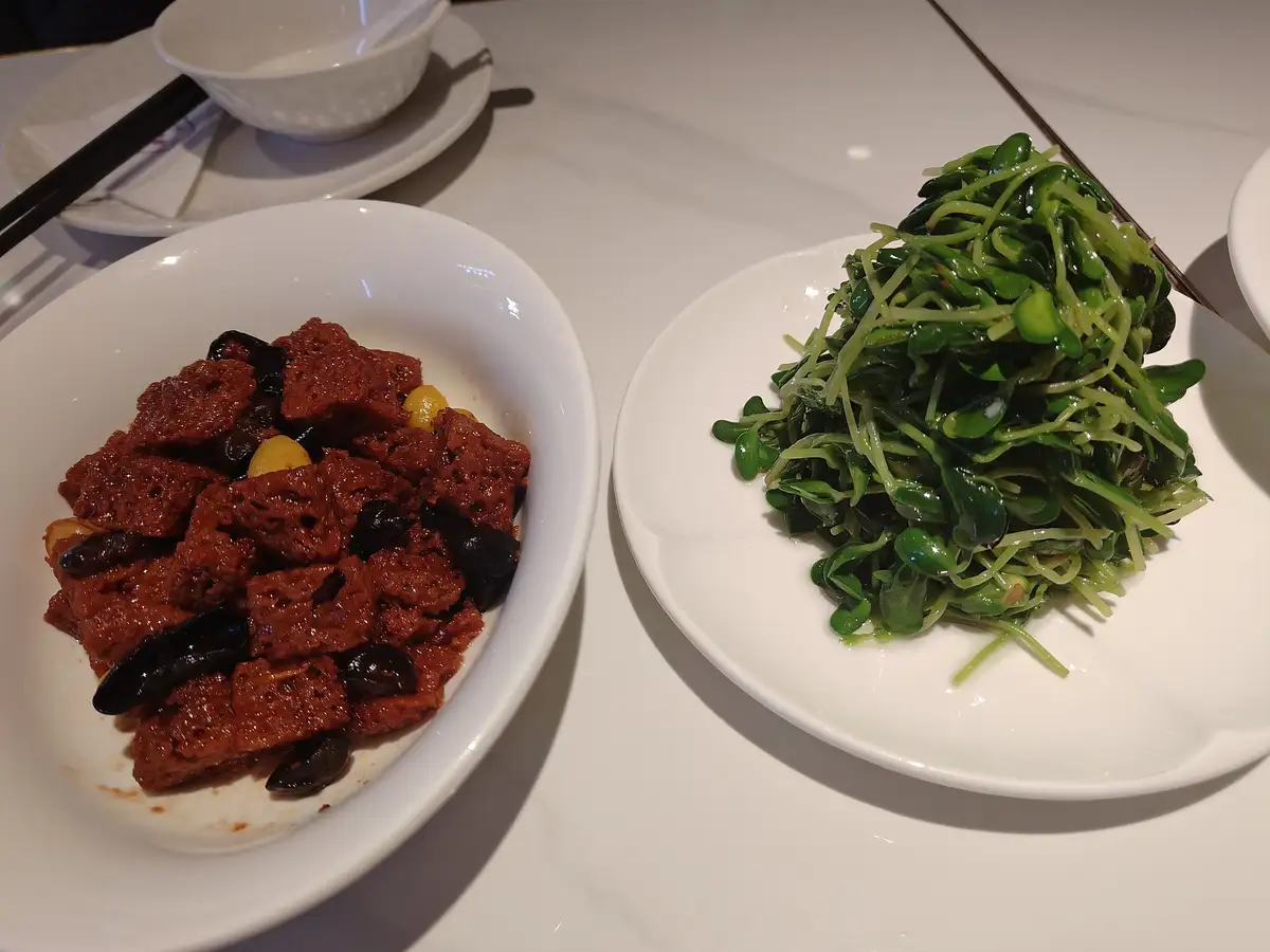 two dishes on a white table: one with reddish-brown cubes and black olives, the other with green sprouts. overlaid text…
