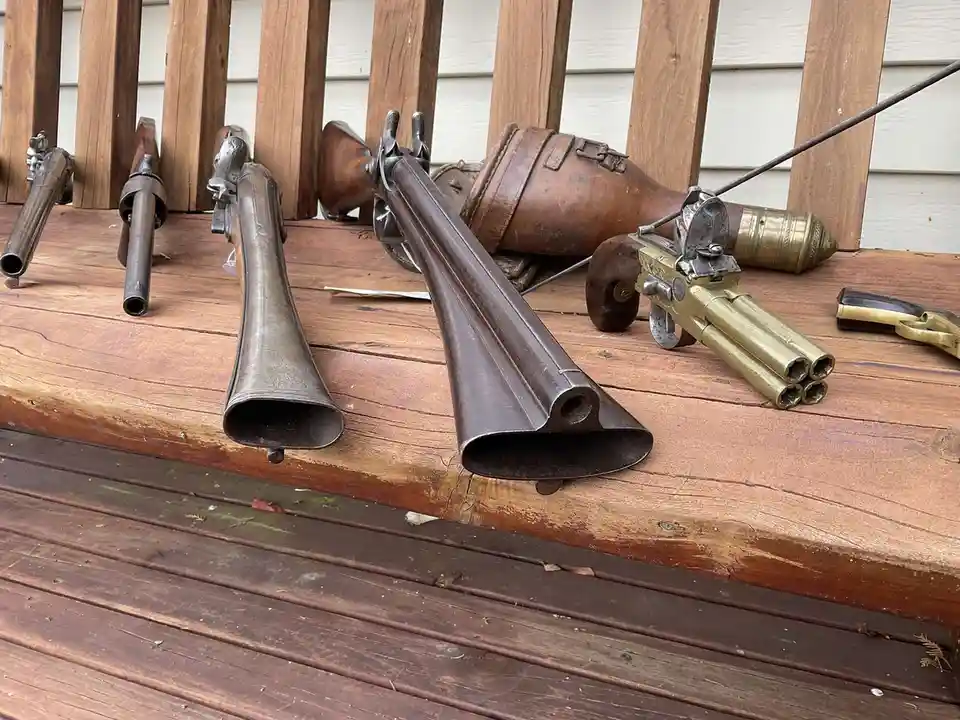 Several antique firearms, including flintlock pistols and a multi-barrelled brass pistol, are arranged on a wooden deck…