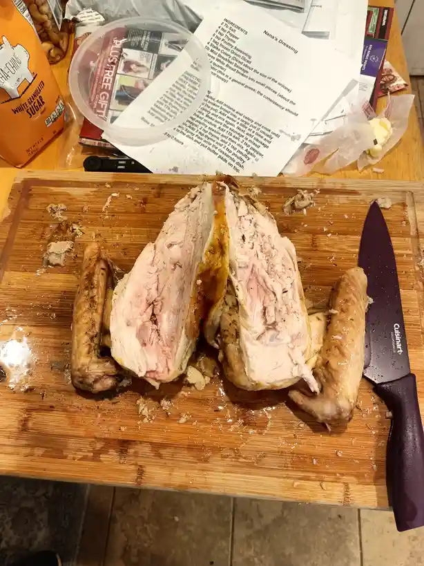 A roasted chicken, split open and sliced, sits on a wooden cutting board next to a purple-handled knife. Behind it, a…