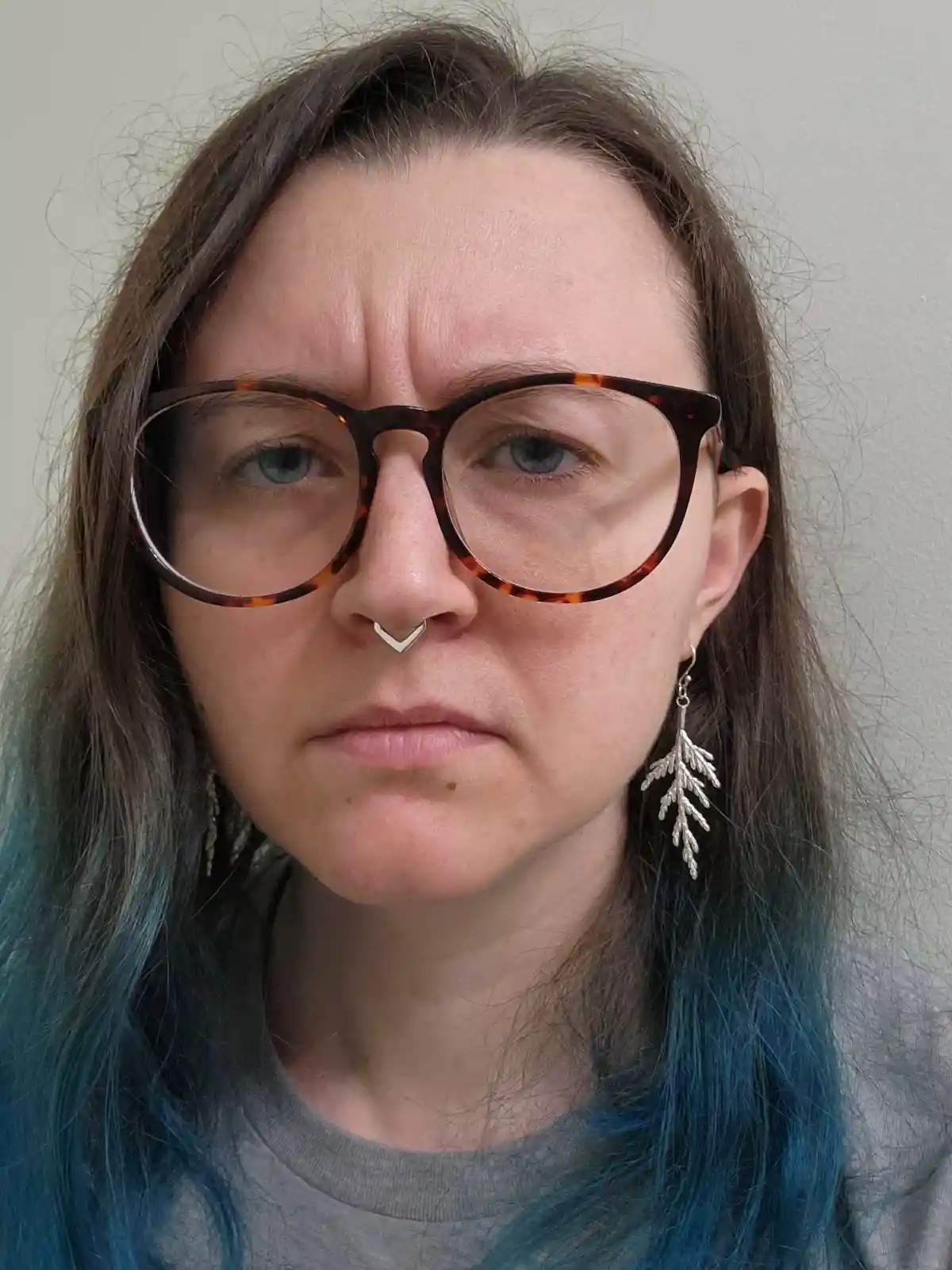 A person with long brown hair dyed blue at the ends, wearing tortoiseshell glasses, a silver nose ring, and leaf-shaped…
