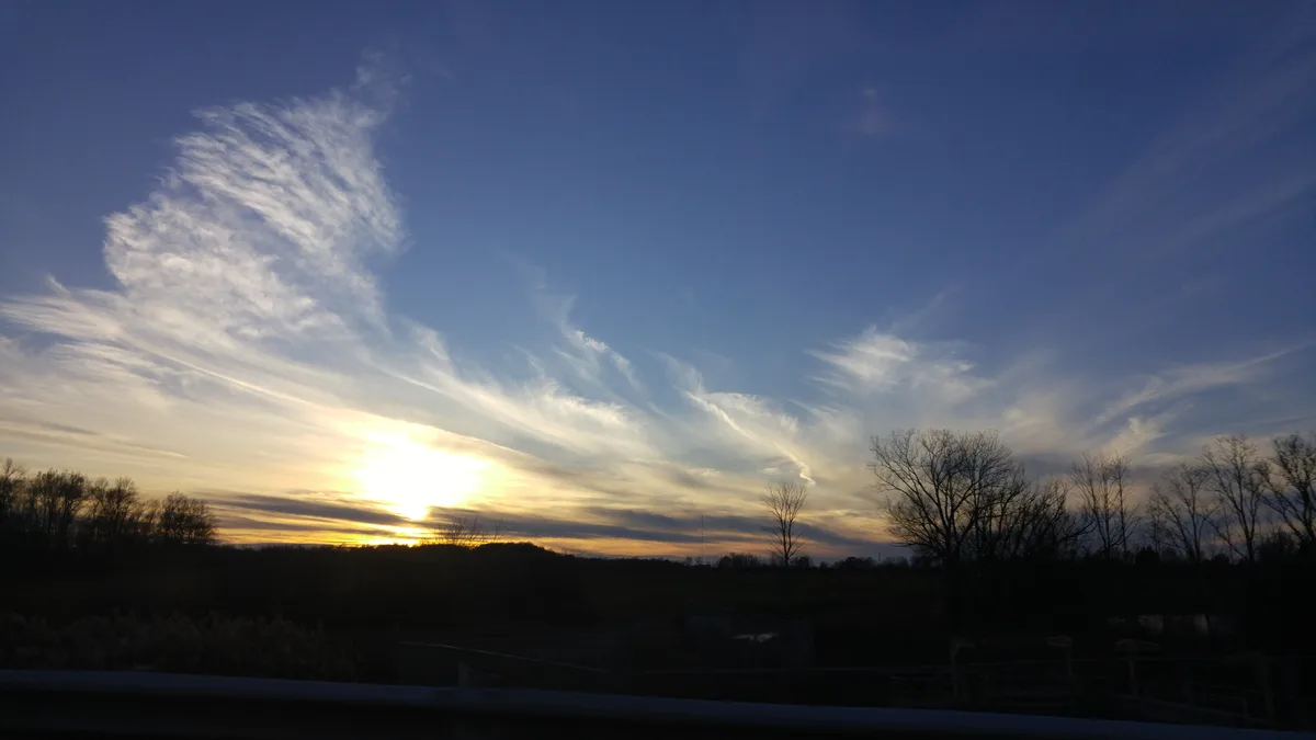 A scenic sunset photo shows a bright sun low on the horizon, casting golden light across wispy clouds against a deep blue…