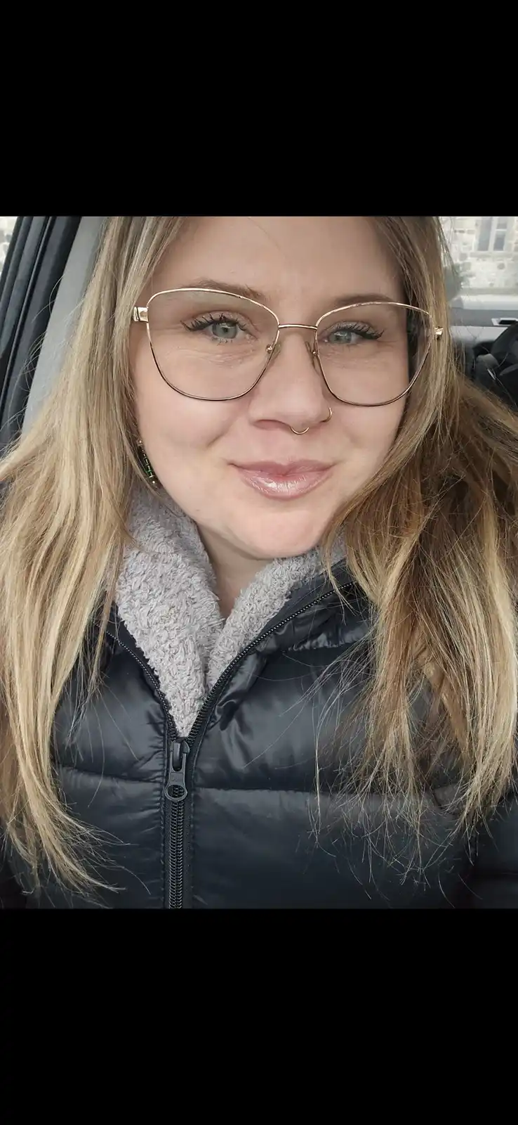 A selfie of a woman with blonde hair, wearing gold-rimmed glasses and a black puffer jacket with a grey fleece collar…