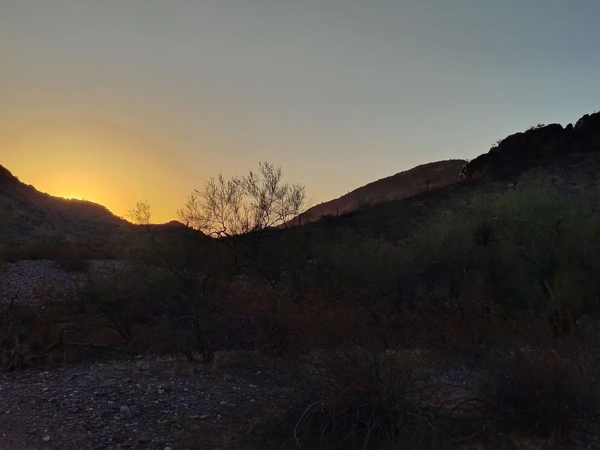 A desert landscape at sunset with silhouetted hills and sparse vegetation under a gradient sky transitioning from orange to…