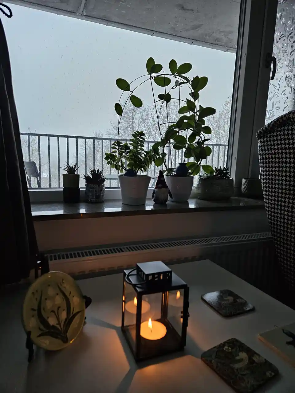 A cozy indoor scene with a lit candle in a lantern on a table, surrounded by decorative items. On the windowsill, several…