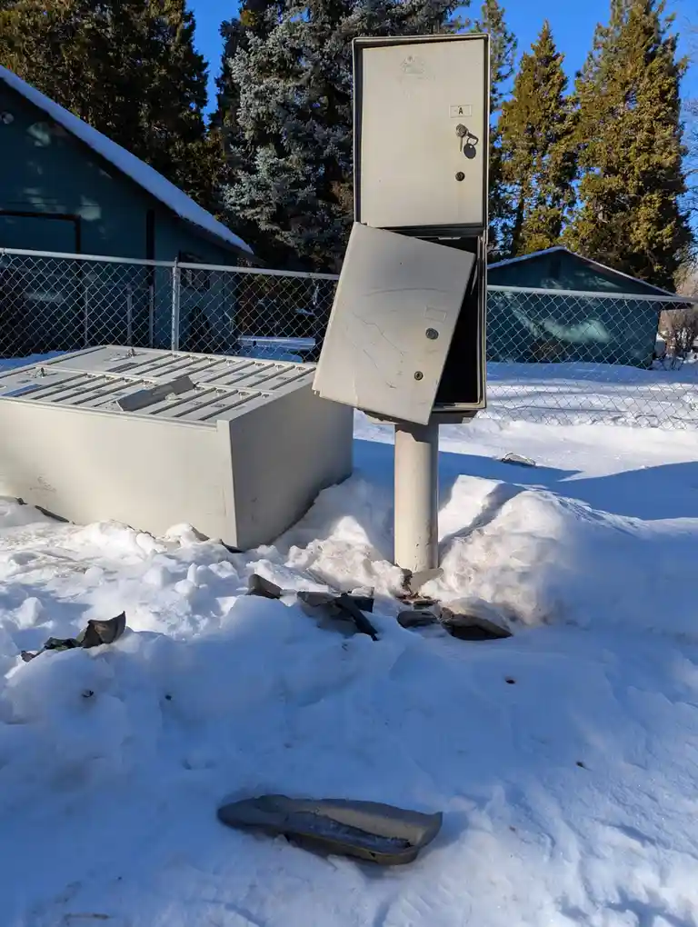 An outdoor utility box with its door ajar stands in deep snow, surrounded by scattered debris and a broken plastic sled. The…
