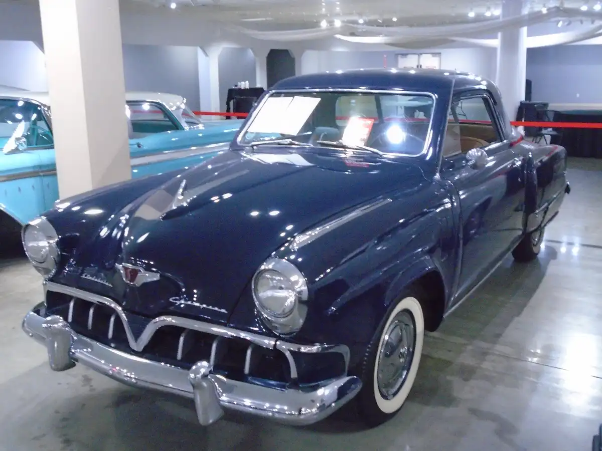 A shiny, dark blue 1950s classic car, likely a Chrysler, is displayed indoors in a museum or showroom with polished floors…