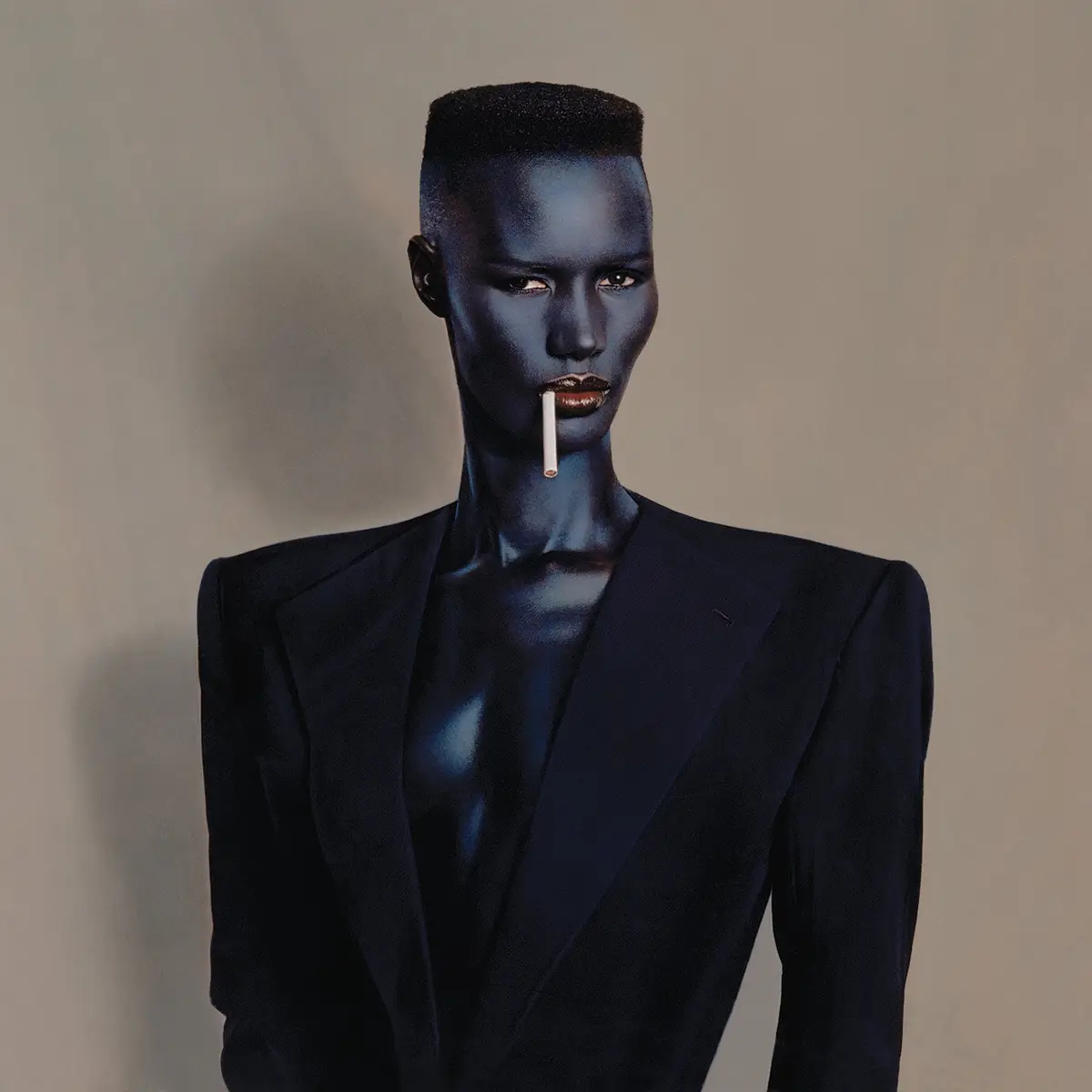 A person with dark blue skin and a short haircut wears a dark suit and holds a cigarette between their lips. The overlaid…